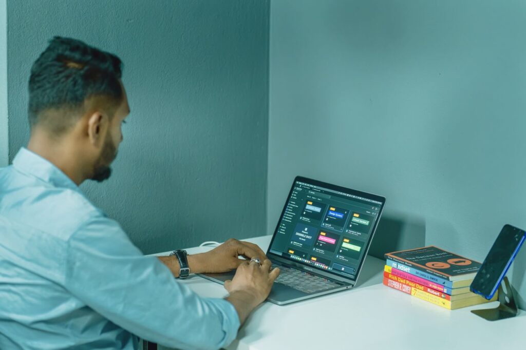 pexels-photo-16323455-16323455 Focused man working on laptop at home office desk, surrounded by books and smartphone for a productive work environment.